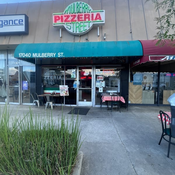 Mulberry Pizzeria Street storefront with a green awning, outdoor seating, and a neon sign.